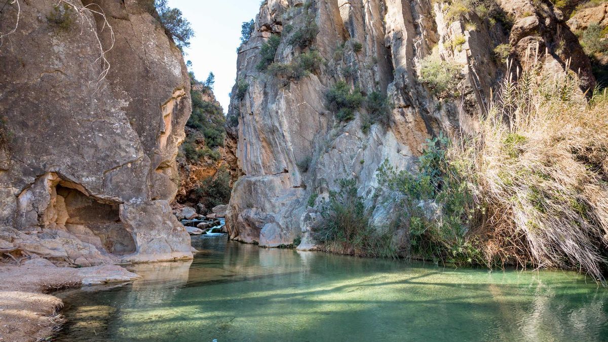 La ciudad medieval con tres monumentos que son Bien de Interés Cultural y paisajes fluviales que es perfecta para recorrer a pie En el corazón de la comarca valenciana de Los Serranos, Chelva invita a caminar entre callejuelas, monumentos históricos y senderos marcados por el rumor del aguaLa ruta de senderismo en València que conecta naturaleza y patrimonio mientras caminas junto al agua
Plazas, senderos, fuentes, miradores y edificios que han ido acumulando siglos de historia. Chelva, en el interior de la provincia de Valencia y rodeada por los paisajes montañosos de la comarca de Los Serranos, pertenece a esa categoría de pueblos que invitan a dejarse llevar por el territorio.
A poco más de una hora de la capital valenciana, esta localidad conserva una de las herencias históricas más interesantes del interior mediterráneo. Pero, más allá de sus calles medievales o de la fama de algunos de sus rincones más fotografiados, Chelva destaca por una combinación menos habitual: la convivencia entre un patrimonio monumental de primer nivel y un paisaje modelado por el agua. Aquí las rutas a pie enlazan antiguos palacios, ermitas, puentes, lavaderos y caminos fluviales que convierten cualquier paseo en una pequeña aventura cultural y natural.
La mejor forma de descubrirla consiste precisamente en eso: caminar sin prisas. Porque Chelva es un lugar para recorrerlo como quien hojea un libro antiguo, deteniéndose en los detalles.
Tres Bienes de Interés Cultural entre montañas y barrancos
Pocas localidades de dimensiones modestas pueden presumir de reunir tres espacios protegidos con la máxima categoría patrimonial. Chelva cuenta con tres elementos declarados Bien de Interés Cultural que ayudan a explicar la importancia histórica que tuvo este territorio a lo largo de los siglos.
El primero es su Conjunto Histórico, un reconocimiento que protege buena parte del casco antiguo y su singular trazado urbano medieval. Más que un monumento concreto, se trata de un paisaje urbano completo donde la historia sigue formando parte de la vida cotidiana.
El segundo es el Palacio Vizcondal, cuya relevancia histórica y arqueológica lo ha convertido en una referencia imprescindible para comprender la evolución de la localidad. Se trata de uno de los edificios más representativos, situado junto a la Plaza Mayor. Su origen se remonta al siglo XIV, cuando fue residencia de los vizcondes de Chelva, aunque las investigaciones arqueológicas han demostrado que bajo sus muros se esconden vestigios mucho más antiguos.
Palacio Vizcondal de Chelva.
El edificio incorpora restos de un alcázar almohade del siglo XII y evidencias de ocupaciones anteriores, lo que convierte al conjunto en una especie de resumen arquitectónico de la evolución histórica de la localidad. Declarado Bien de Interés Cultural, constituye una de las piezas patrimoniales más importantes de toda la comarca.
Y el tercero se encuentra fuera del núcleo urbano, integrado en el paisaje de barrancos y montañas que rodean Chelva. Se trata del acueducto romano de Peña Cortada, una de las obras de ingeniería hidráulica más espectaculares conservadas en España. Construido durante el siglo I d. C., todavía mantiene visibles algunos de sus tramos más impresionantes, incluidos puentes, galerías excavadas en la roca y pasos elevados enclavados en el municipio vecino de Calles y que continúan asombrando a quienes los recorren dos mil años después.
Tramo del acueducto romano de Peña Cortada, en el municipio de Calles, muy próximo a Chelva.
La magnitud de esta infraestructura resulta aún más sorprendente cuando aparece de forma repentina entre pinares, laderas y cortados de roca. No se visita únicamente como monumento: se experimenta caminando.
Otro de los símbolos de Chelva es la iglesia arciprestal de Nuestra Señora de los Ángeles. Considerada una de las mejores muestras del barroco valenciano en el interior de la provincia, su silueta domina el casco urbano y sirve de referencia para orientarse durante cualquier paseo por la población.
Además, el patrimonio local va mucho más allá de sus edificios principales. Antiguos lavaderos, restos de murallas, ermitas dispersas por el término municipal, puentes históricos y construcciones tradicionales forman parte de un conjunto que convierte la visita en una experiencia continua de descubrimiento.
El agua como compañera de viaje
Si el patrimonio explica el pasado de Chelva, el agua define su carácter. El municipio está rodeado por un entorno natural donde manantiales, fuentes, acequias y cursos fluviales han condicionado la vida de sus habitantes durante generaciones.
Las rutas senderistas que recorren el término municipal permiten descubrir una geografía especialmente amable para quienes disfrutan caminando. Los caminos avanzan entre bosques mediterráneos, terrazas agrícolas tradicionales y rincones donde el agua aparece constantemente, ya sea brotando de una fuente histórica o acompañando el recorrido de antiguos sistemas hidráulicos.
Esa presencia permanente genera una sensación poco habitual en el interior valenciano. Incluso durante los meses más cálidos, muchos de estos itinerarios conservan áreas de sombra y pequeños oasis naturales que invitan a detenerse.
Entre todas las propuestas destaca el recorrido vinculado precisamente al acueducto de Peña Cortada. La ruta permite acercarse a esta obra que representa uno de los mayores logros de la ingeniería romana en la Comunitat Valenciana. A medida que el sendero gana altura, aparecen puentes suspendidos sobre barrancos, galerías excavadas directamente en la roca y miradores naturales desde los que contemplar el paisaje serrano.
Lo más llamativo es que naturaleza e historia parecen inseparables. El monumento no se entiende sin el territorio y el territorio adquiere una nueva dimensión gracias a la presencia del monumento.
Quizá ahí resida el verdadero atractivo de Chelva. En su capacidad para ofrecer una experiencia completa sin necesidad de artificios. Aquí el viajero puede comenzar la mañana recorriendo plazas históricas, descubrir siglos de historia en un palacio medieval, seguir el sonido del agua por senderos fluviales y terminar la jornada contemplando cómo las montañas de Los Serranos se tiñen de tonos dorados al atardecer.
Ruta por los Barrios Históricos
Y aunque Chelva es uno de esos destinos que invitan a dejarse llevar por la intuición, una buena forma de comprender la complejidad de su pasado es sumarse a alguna de las visitas guiadas que se organizan durante todo el año. La ruta por los Barrios Históricos permite recorrer durante unas dos horas los distintos espacios que conforman su Conjunto Histórico, desde el antiguo barrio andalusí de Benacacira hasta el Arrabal morisco, pasando por los sectores cristianos y judíos, mientras se descubren episodios, curiosidades y detalles que a menudo pasan desapercibidos si no es gracias a un guía local. Una experiencia accesible y muy recomendable para completar la escapada y entender por qué este rincón de Los Serranos sigue siendo uno de los lugares con más personalidad del interior valenciano.
